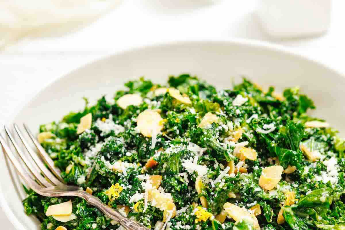 A white bowl filled with kale salad, topped with sliced almonds and grated cheese, with a fork resting on the side.
