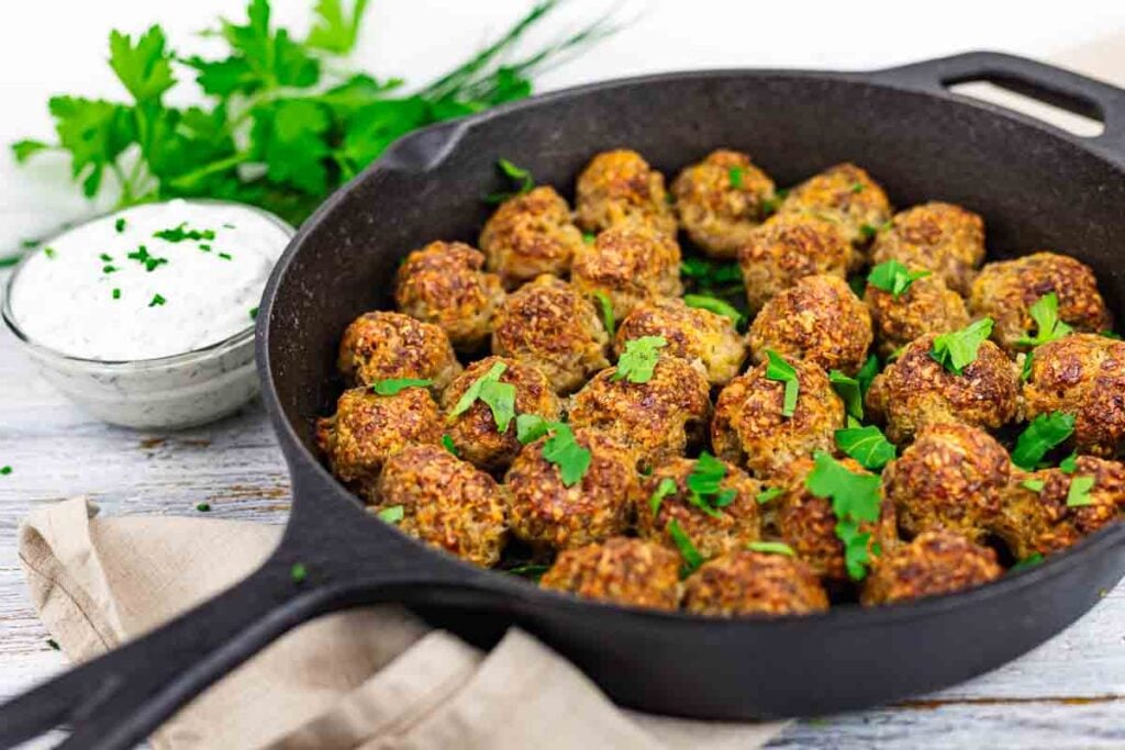 A cast iron skillet filled with cooked meatballs garnished with herbs, with a bowl of creamy dipping sauce and fresh parsley in the background.