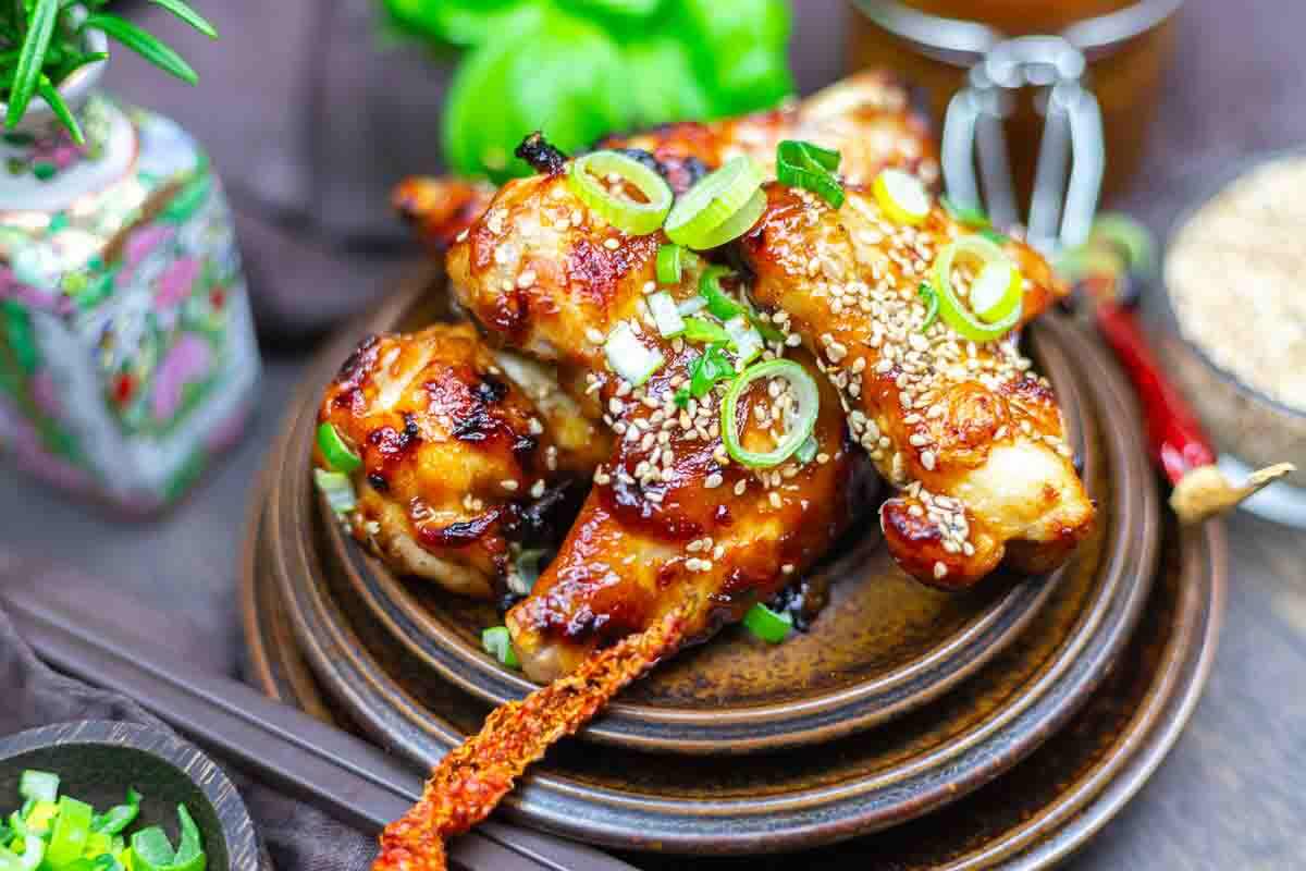 A plate of glazed chicken drumsticks garnished with sesame seeds and chopped green onions, with a side of red chili pepper and chopsticks nearby.