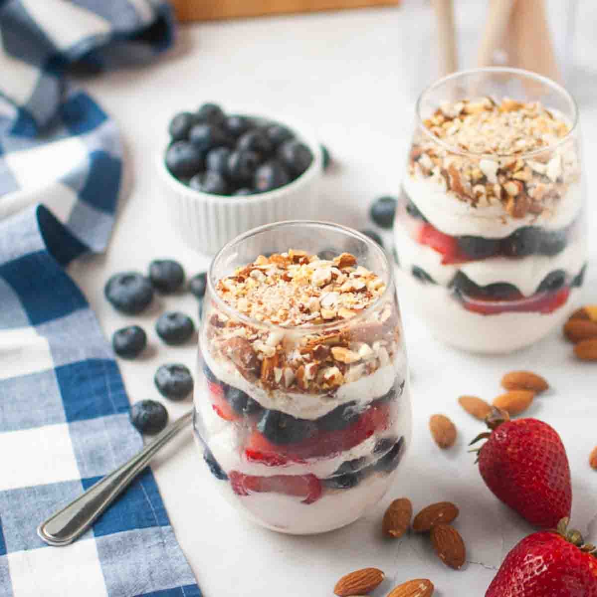 Two glasses of layered parfait with yogurt, strawberries, blueberries, and granola, surrounded by almonds, blueberries, and strawberries on a white surface.