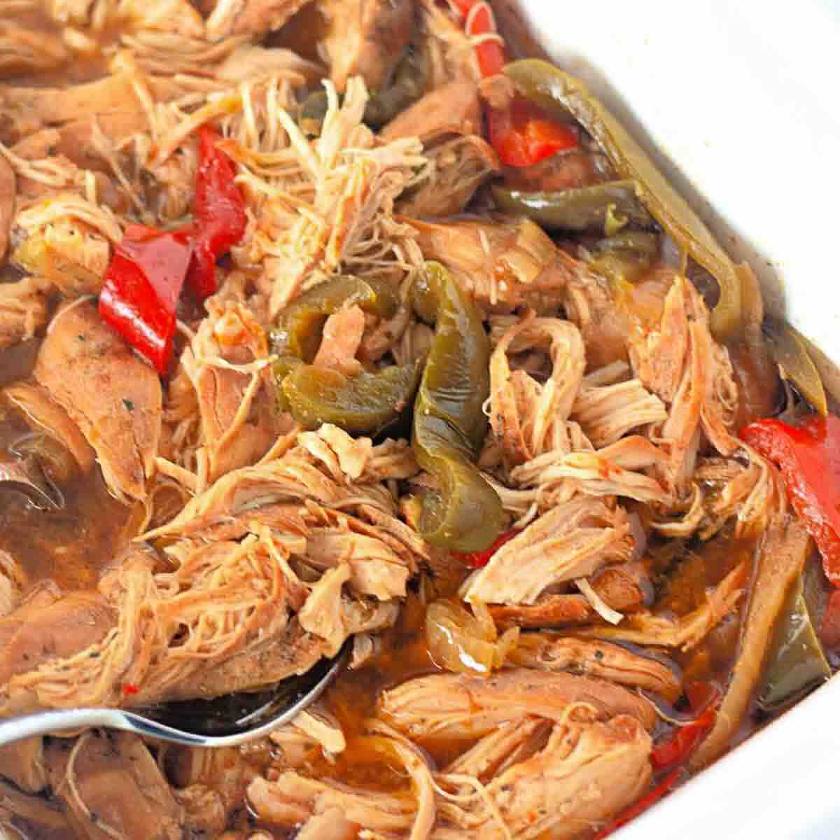 Shredded cooked chicken with red and green bell peppers in a saucy mixture, served in a white dish with a spoon.