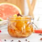 A glass jar filled with spiced watermelon rind pickles sits on a white surface, with a slice of watermelon and whole spices in the background.