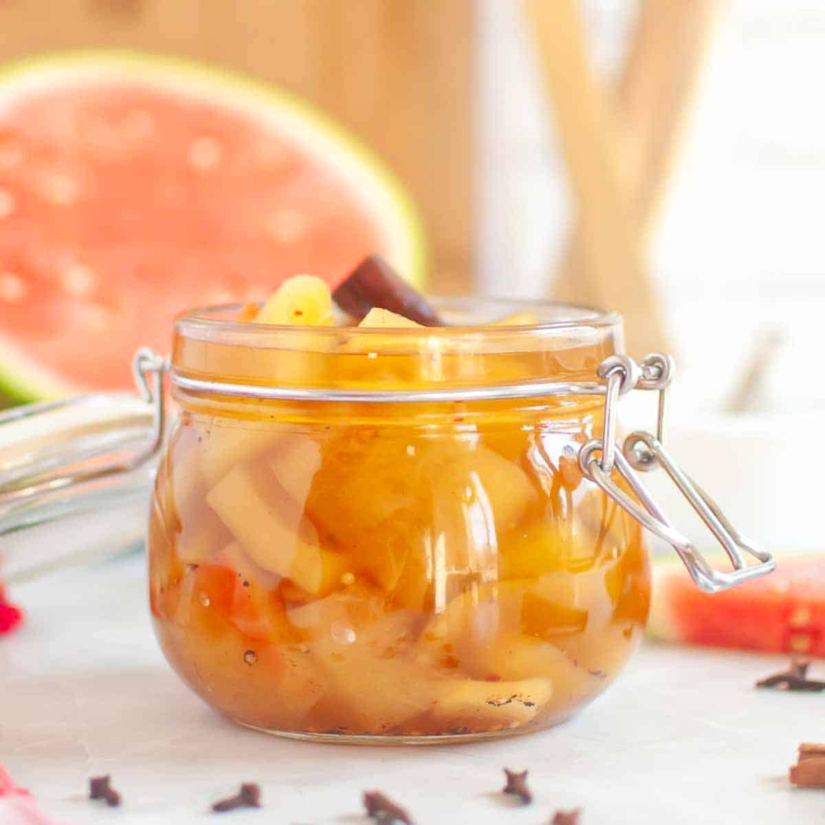 A glass jar filled with spiced watermelon rind pickles sits on a white surface, with a slice of watermelon and whole spices in the background.