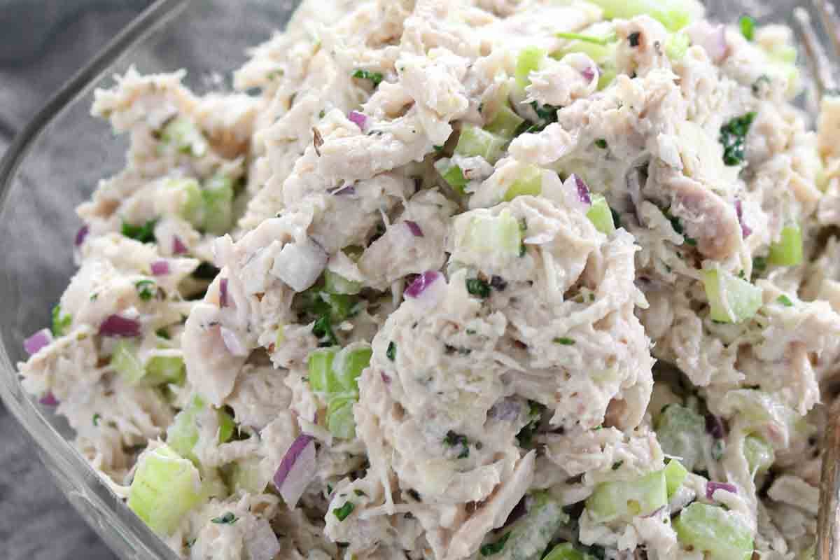 Close-up of chicken salad with shredded chicken, chopped celery, red onion, and herbs mixed together in a clear glass bowl.
