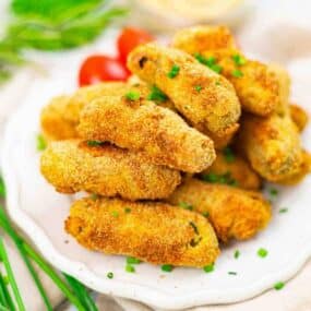 A plate of golden-brown, breaded croquettes garnished with chopped chives, with cherry tomatoes and greens in the background.