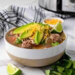 A bowl of soup with meatballs, sliced avocado, shredded cheese, cilantro, and a lime wedge, with a spoon and a striped napkin beside it.