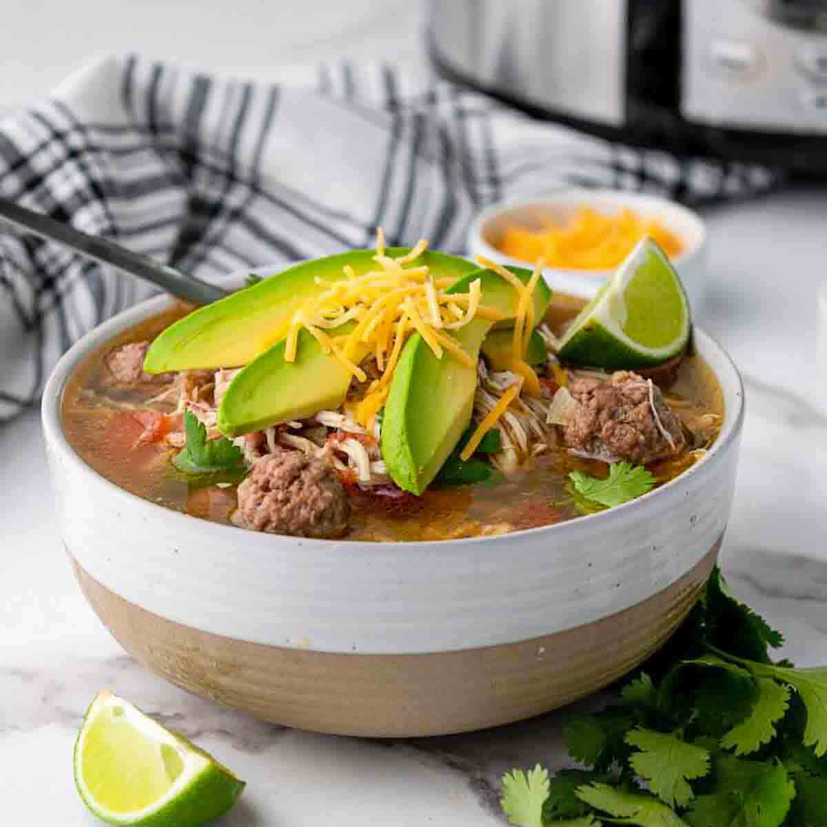 A bowl of soup with meatballs, sliced avocado, shredded cheese, cilantro, and a lime wedge, with a spoon and a striped napkin beside it.
