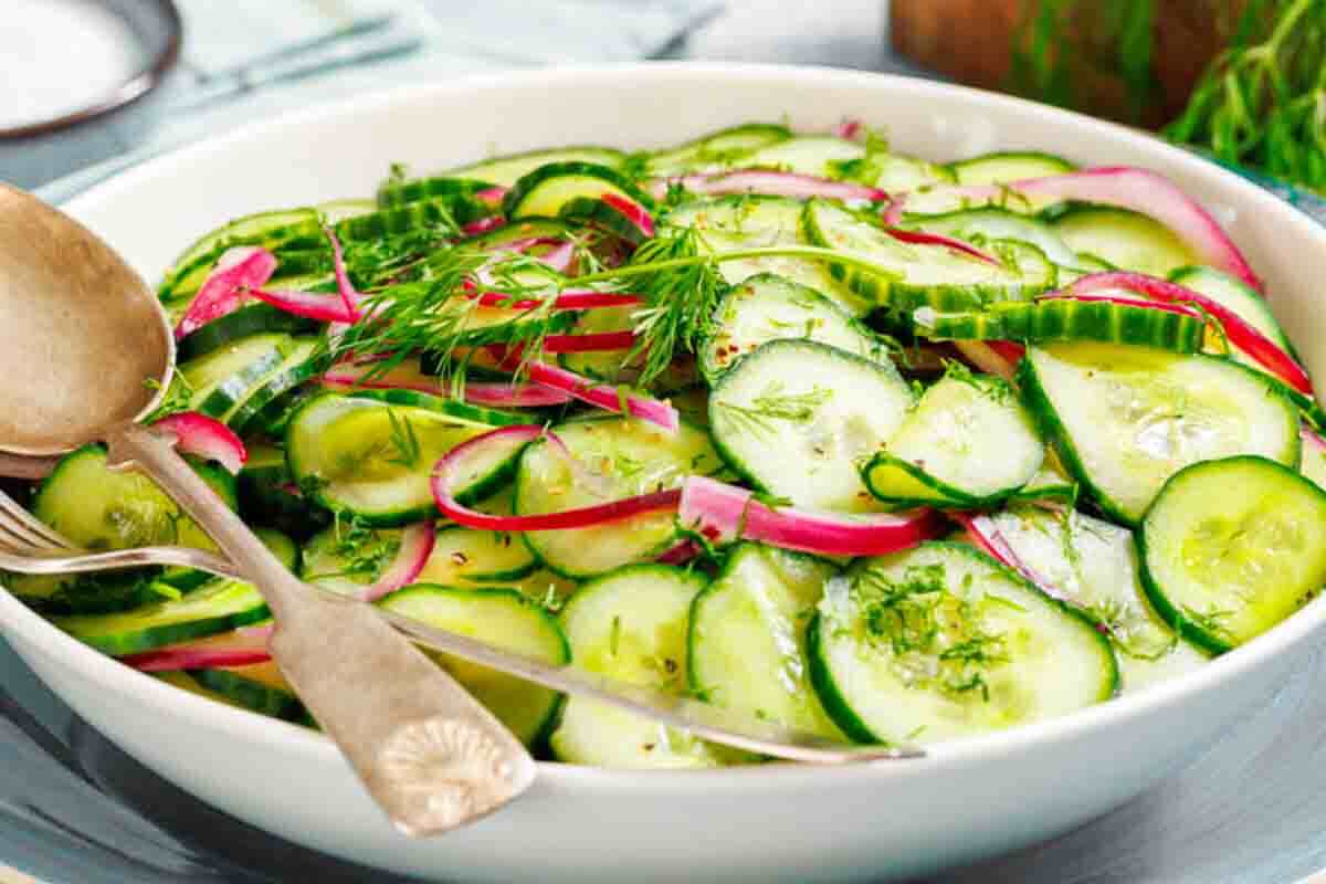 A white bowl filled with cucumber salad, garnished with fresh dill and thin slices of red onion, with serving utensils resting on the side.
