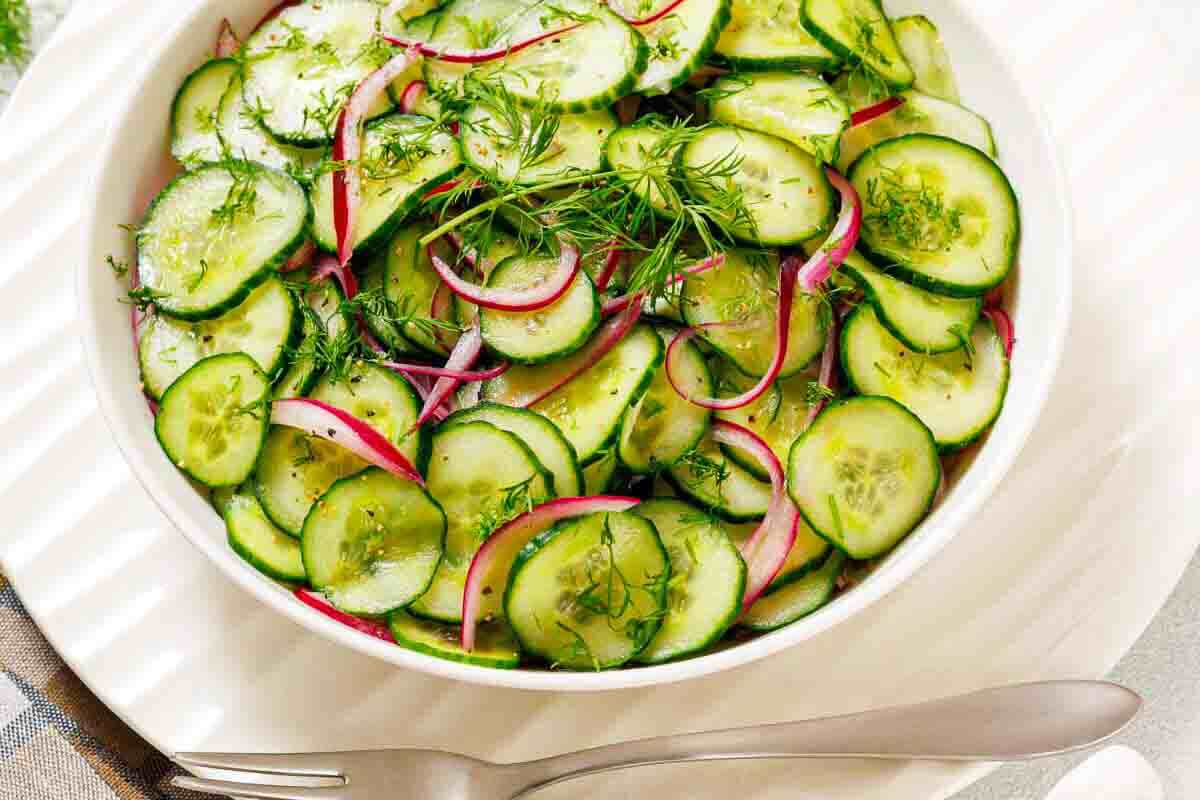A white bowl filled with sliced cucumbers, red onions, and fresh dill, placed on a white plate with a fork and spoon beside it.