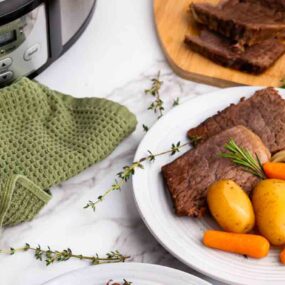 A plate with sliced pot roast, potatoes, and carrots sits on a marble surface near a green towel and a slow cooker.