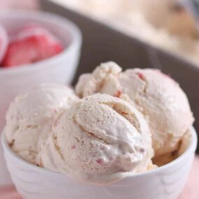 Three scoops of strawberry ice cream are served in a white bowl, with a bowl of frozen strawberries and an ice cream container in the background.
