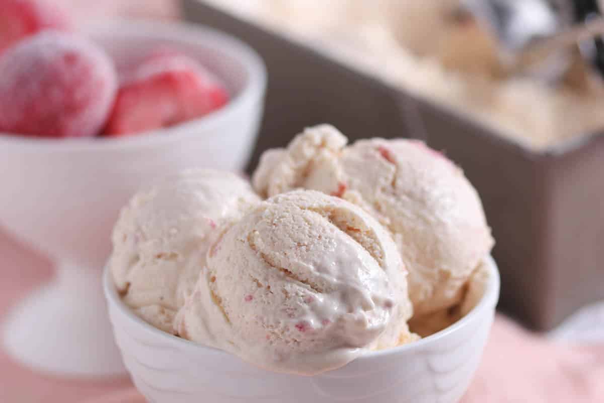 Three scoops of strawberry ice cream are served in a white bowl, with a bowl of frozen strawberries and an ice cream container in the background.