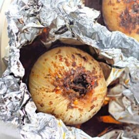 A whole onion seasoned with spices is baked in aluminum foil, showing a browned top and some seasoning on the surface.