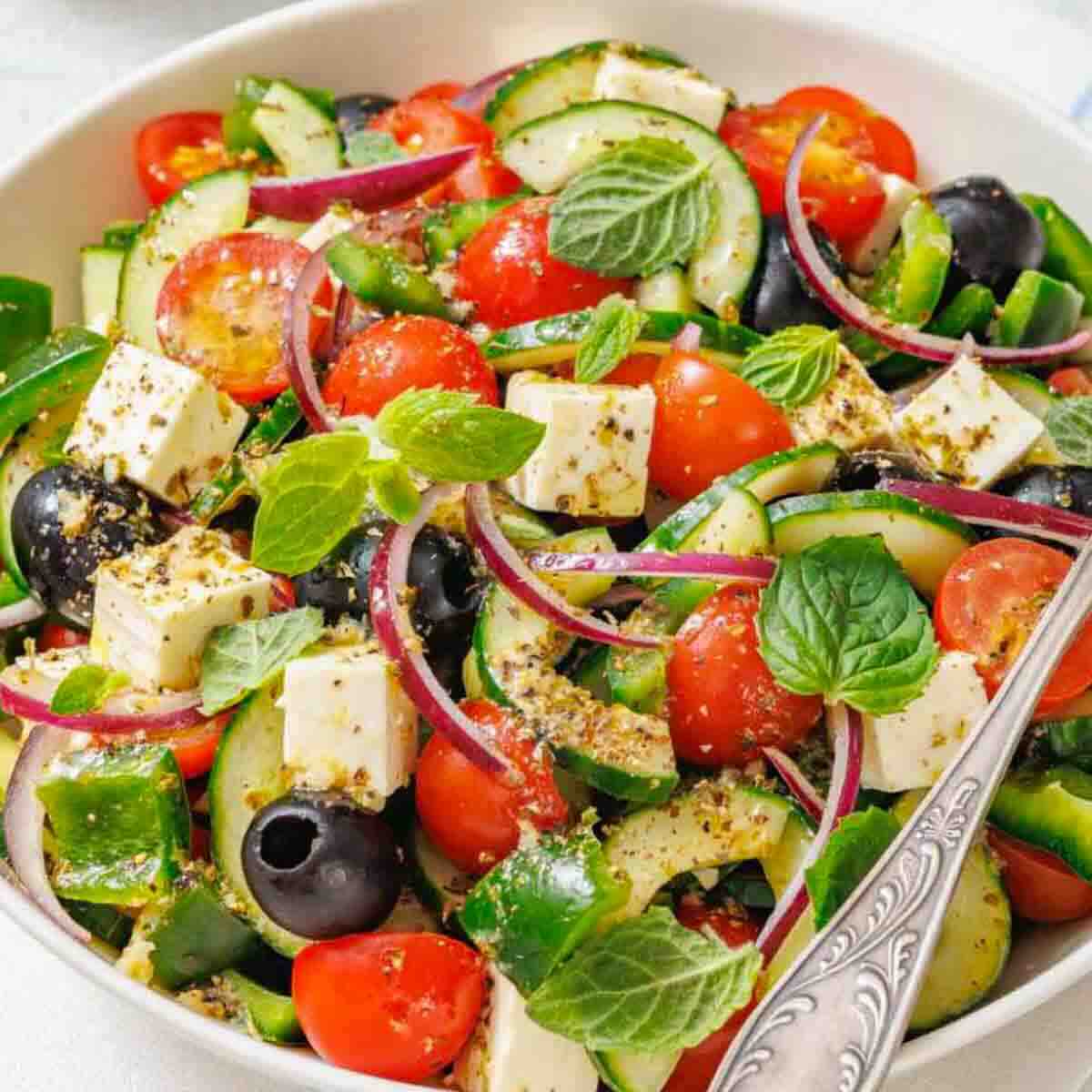 A bowl of Greek salad with cherry tomatoes, cucumber, feta cheese, black olives, red onion, green bell pepper, and fresh mint leaves.