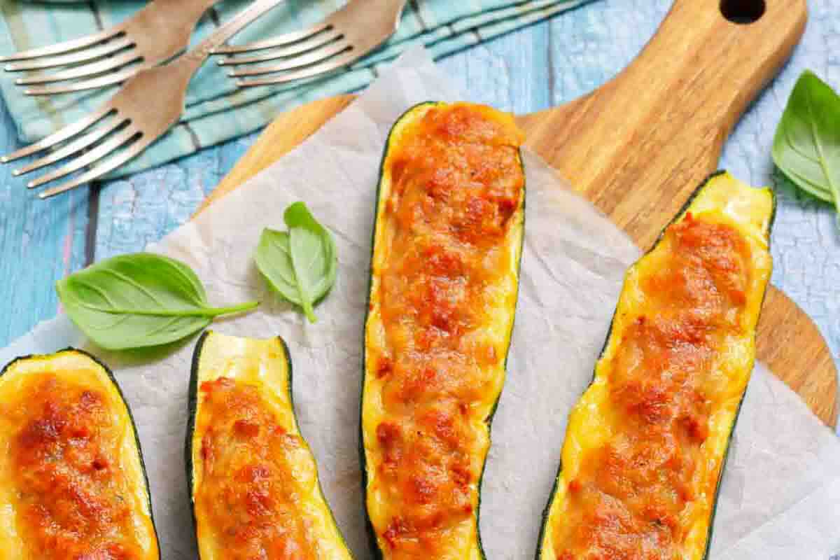 Four baked zucchini halves stuffed with a golden, melted cheese topping are arranged on parchment paper atop a wooden cutting board, with fresh basil leaves nearby.
