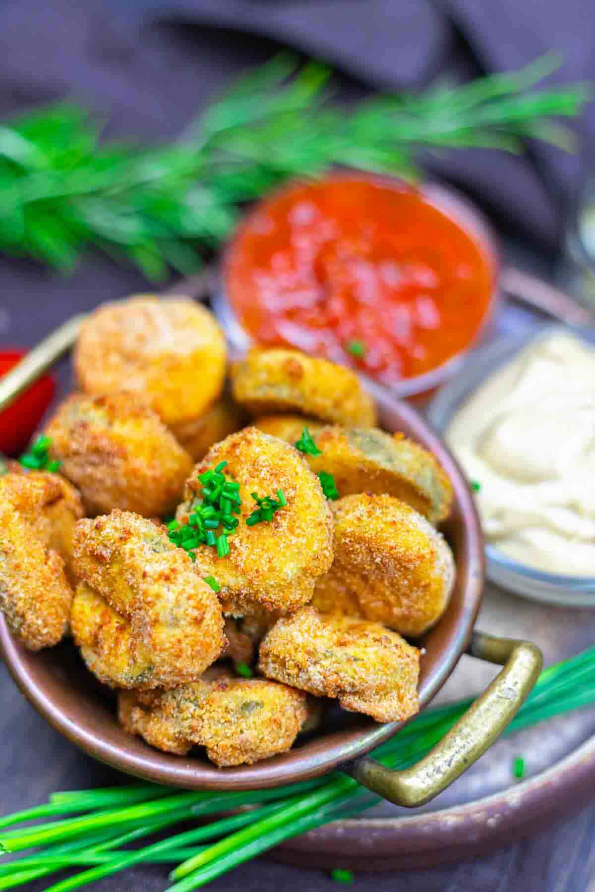 A bowl of breaded and fried mushrooms garnished with chopped chives, served with two dipping sauces in small bowls.