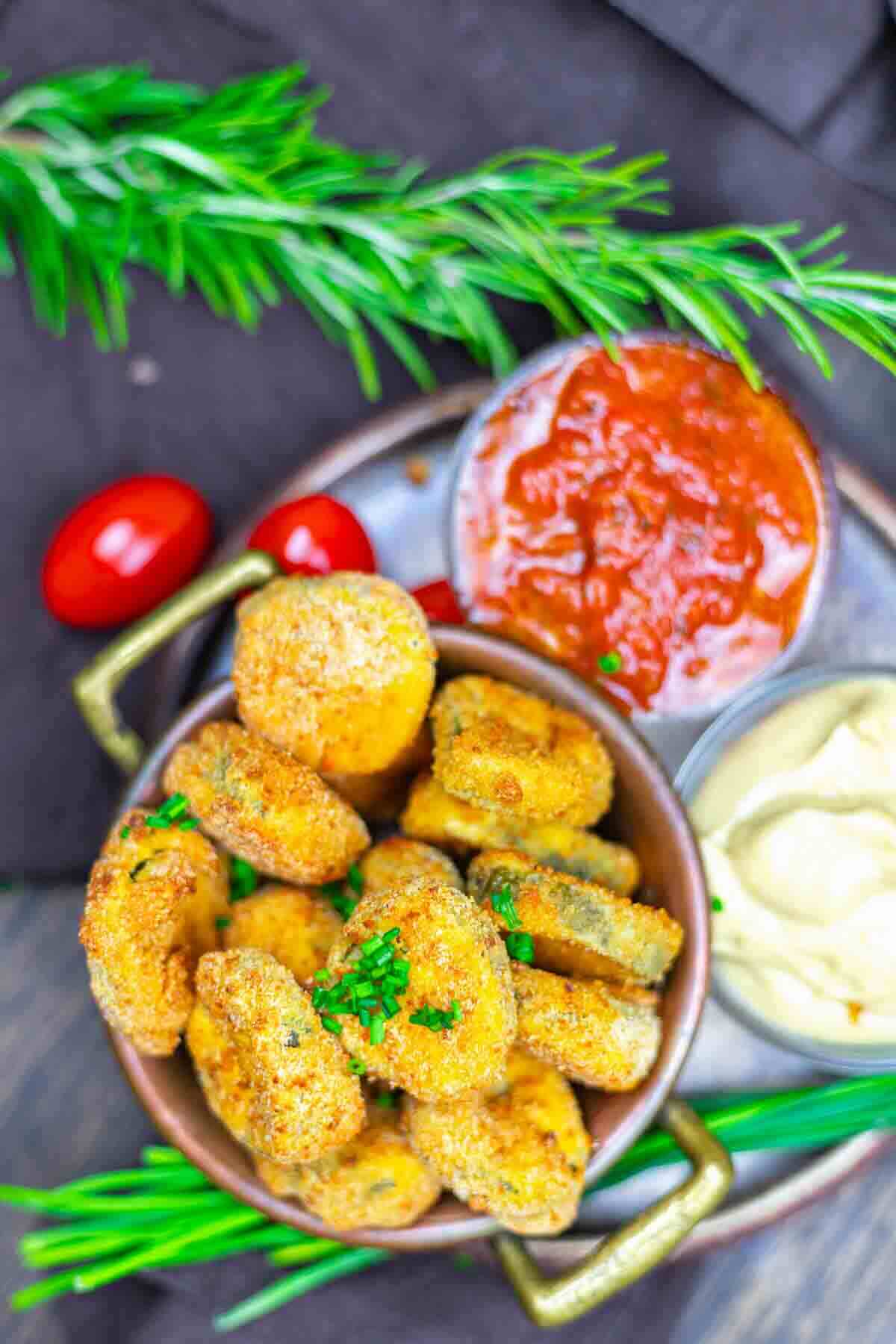 A bowl of breaded fried pickles garnished with chives, served with marinara sauce and a creamy dip, with cherry tomatoes and fresh herbs nearby.