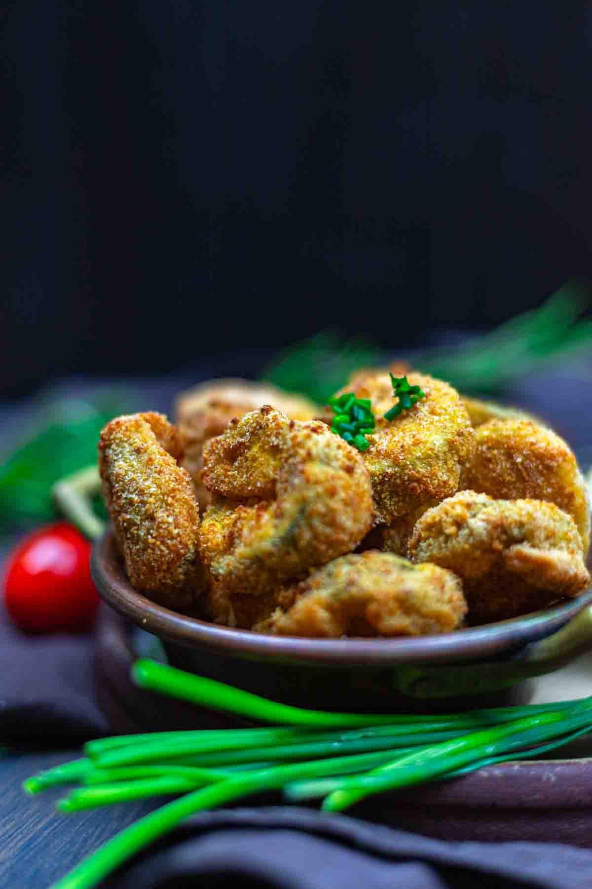 A bowl of breaded and fried mushroom slices garnished with chopped herbs, with green chives and a cherry tomato in the background.
