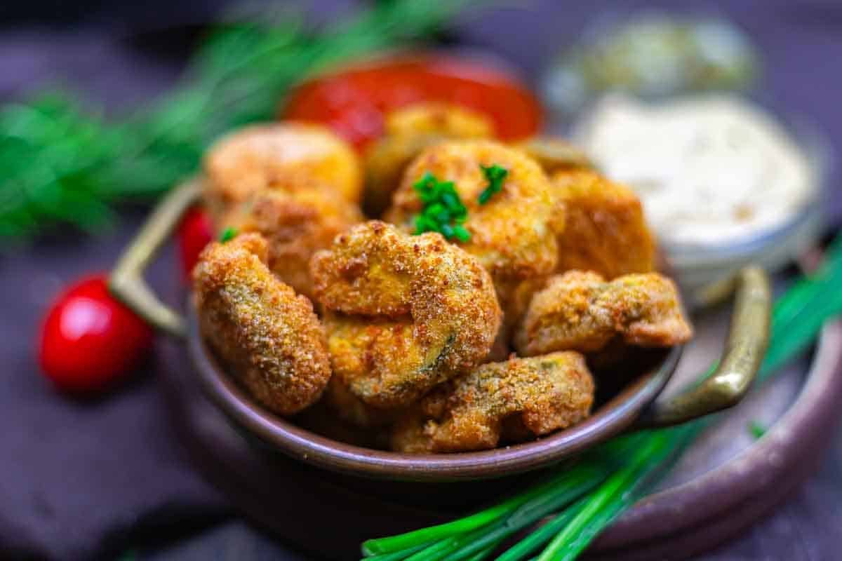 A bowl of breaded and fried pickle pieces garnished with parsley, with dipping sauces and herbs in the background.