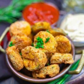 A bowl of breaded, fried pickles garnished with chopped chives, with dipping sauces in the background.