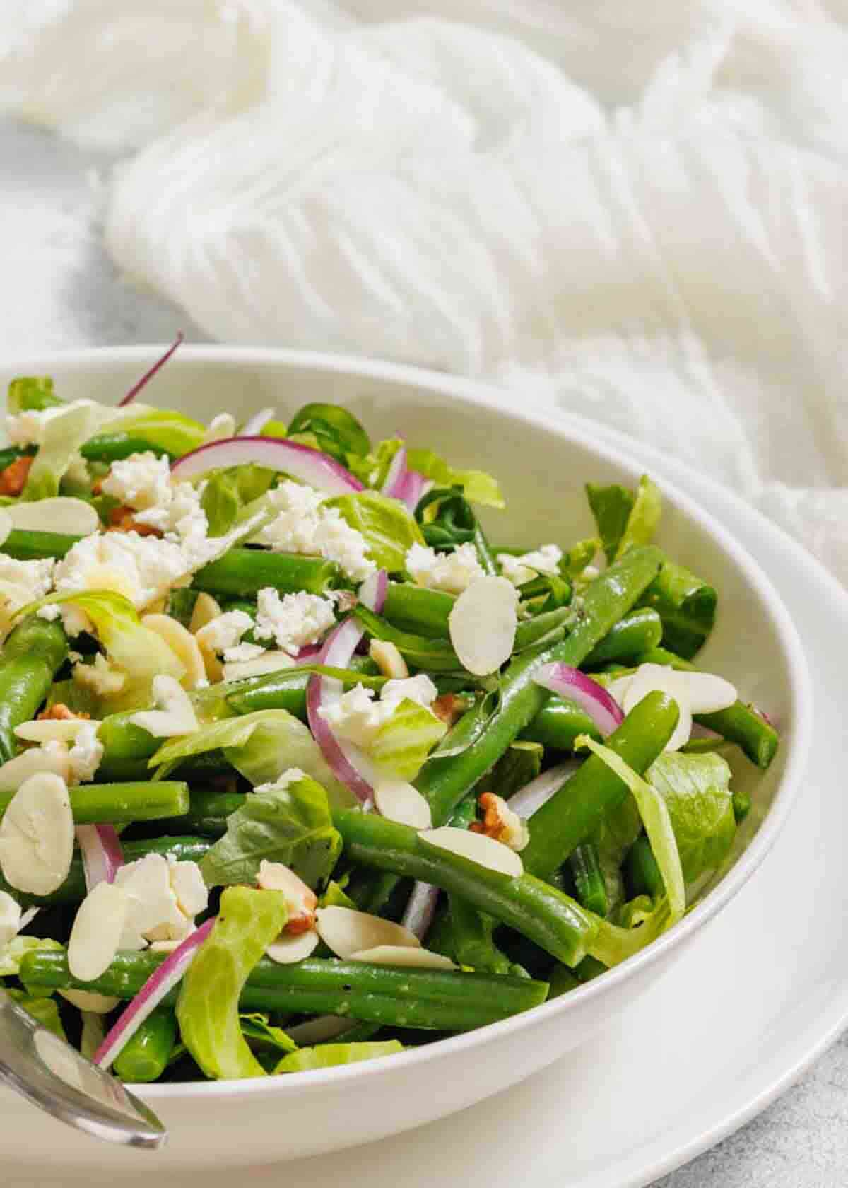 A bowl of salad with green beans, red onion slices, crumbled cheese, almond slices, and lettuce on a white plate.