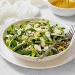 A bowl of green bean salad with feta cheese, red onions, and sliced almonds, served with a fork and a bowl of dressing in the background.