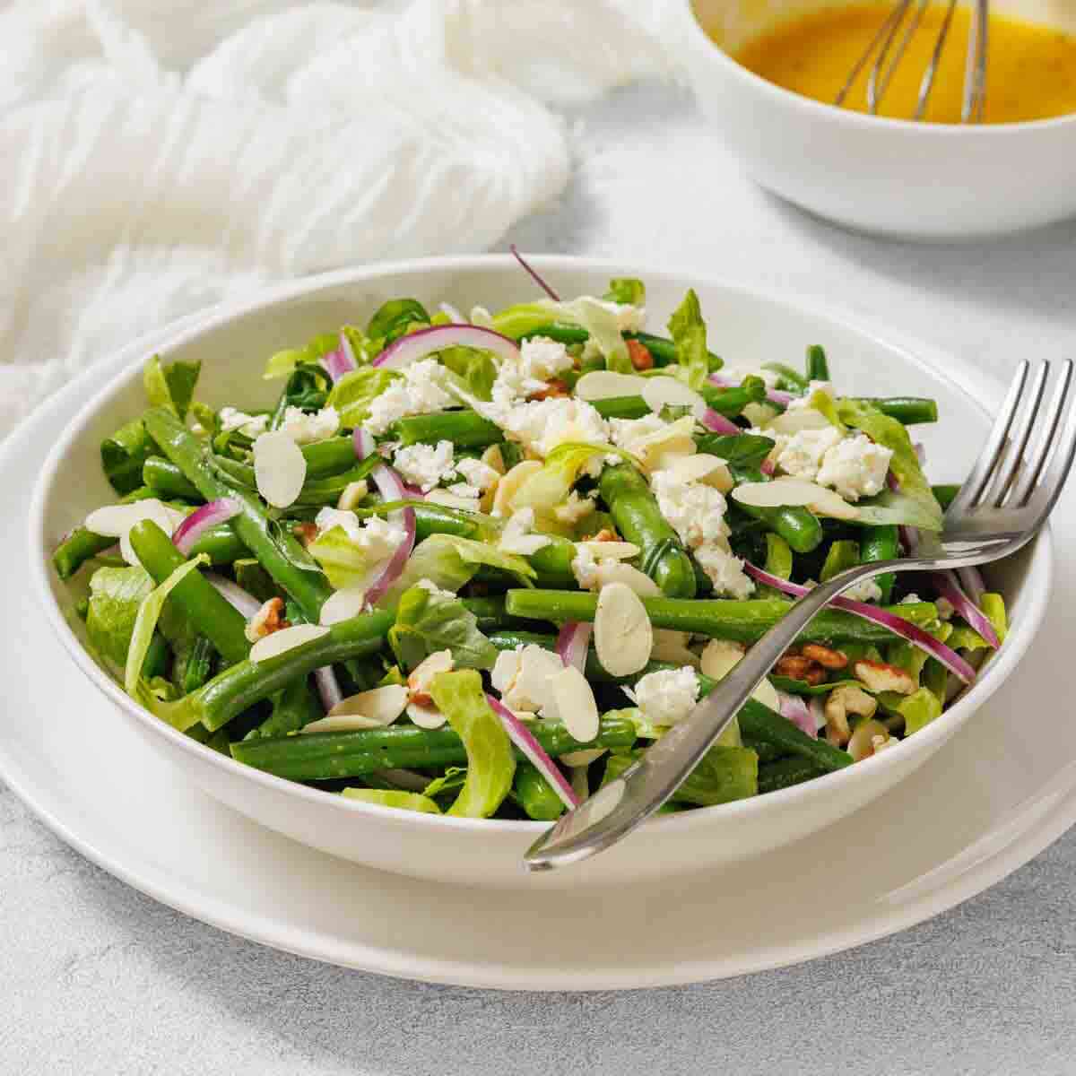 A bowl of green bean salad with feta cheese, red onions, and sliced almonds, served with a fork and a bowl of dressing in the background.