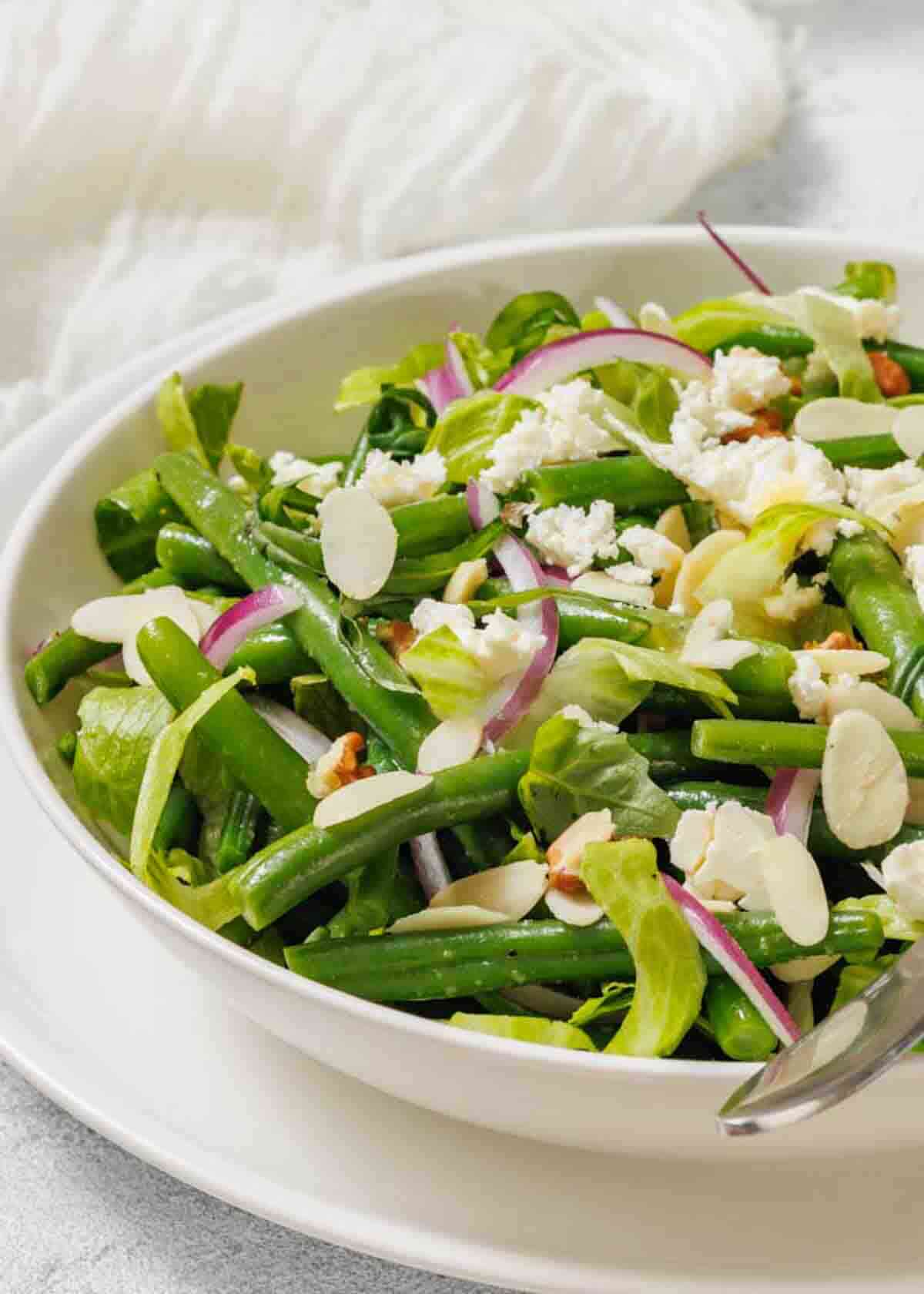 A bowl of green bean salad with sliced almonds, red onion, feta cheese, and mixed greens, served with a metal fork.