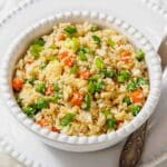 A white bowl filled with cauliflower rice mixed with peas, diced carrots, and chopped green onions, placed on a white plate with a spoon.