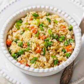 A white bowl filled with cauliflower rice mixed with peas, diced carrots, and chopped green onions, placed on a white plate with a spoon.