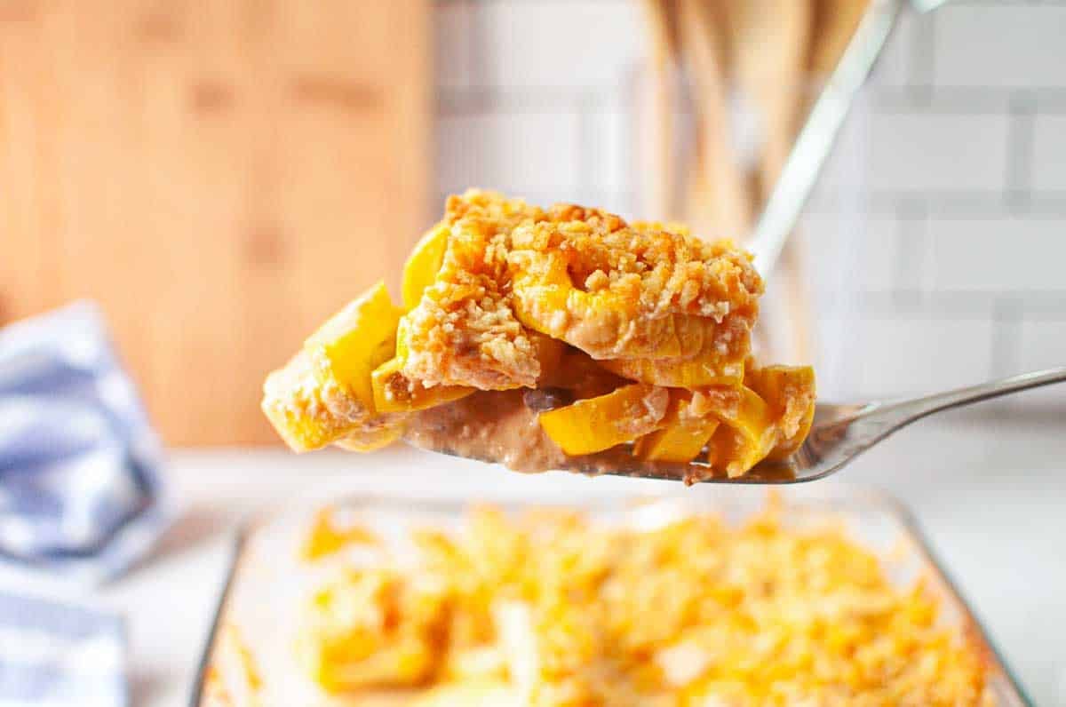 A close-up of a serving spoon holding a portion of cheesy baked casserole with yellow squash and a crumb topping, above a glass baking dish with more casserole.