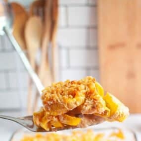 A close-up of a serving spoon holding a piece of peach cobbler with a crumbly topping, kitchen utensils and a wooden cutting board in the background.
