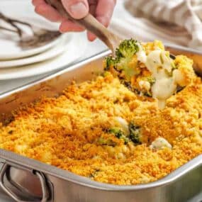 A hand scoops a serving of broccoli and cheese casserole with a golden breadcrumb topping from a rectangular metal baking dish.