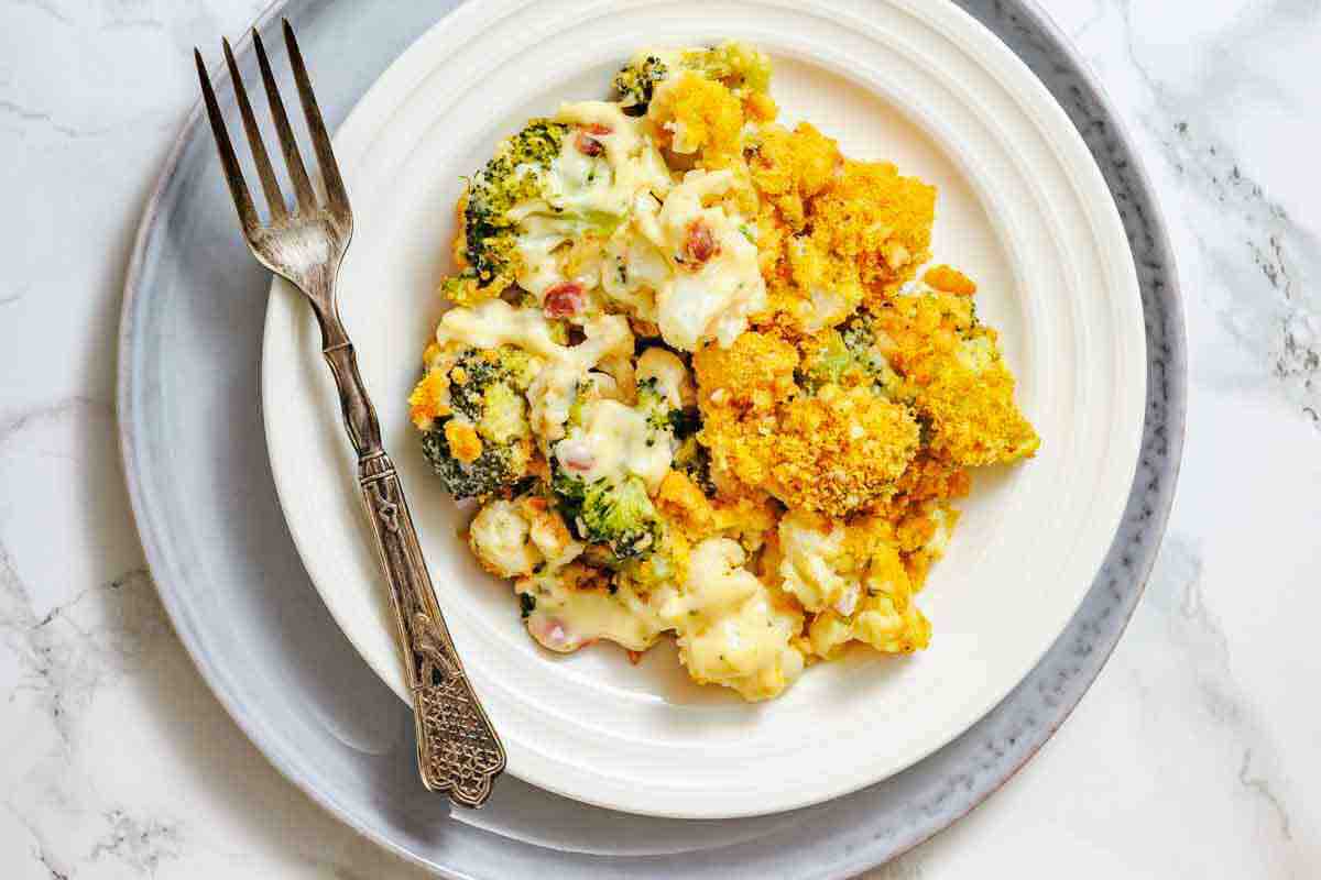 A plate with broccoli, cauliflower, bacon, and cheese casserole topped with breadcrumbs, next to a fork on a marble surface.