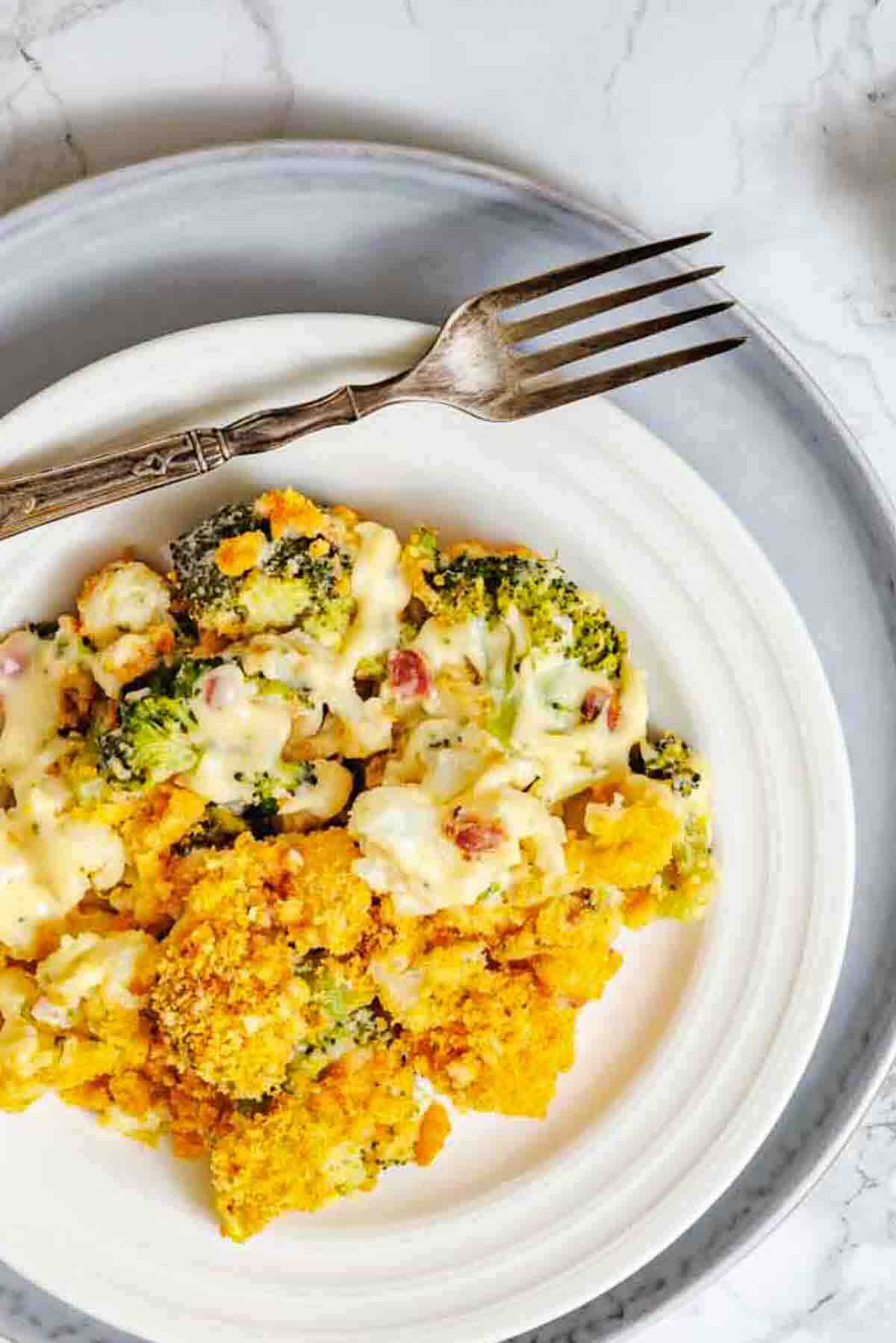 A serving of broccoli casserole with a creamy sauce and crispy topping on a white plate, next to a fork on a marble surface.