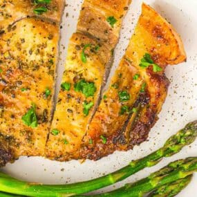 A cooked pork chop garnished with herbs, sliced on a plate with two spears of asparagus.