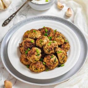 A white plate with a stack of cooked meatballs garnished with chopped green herbs, set on a gray charger plate. Garlic cloves and a bowl of dip are nearby.