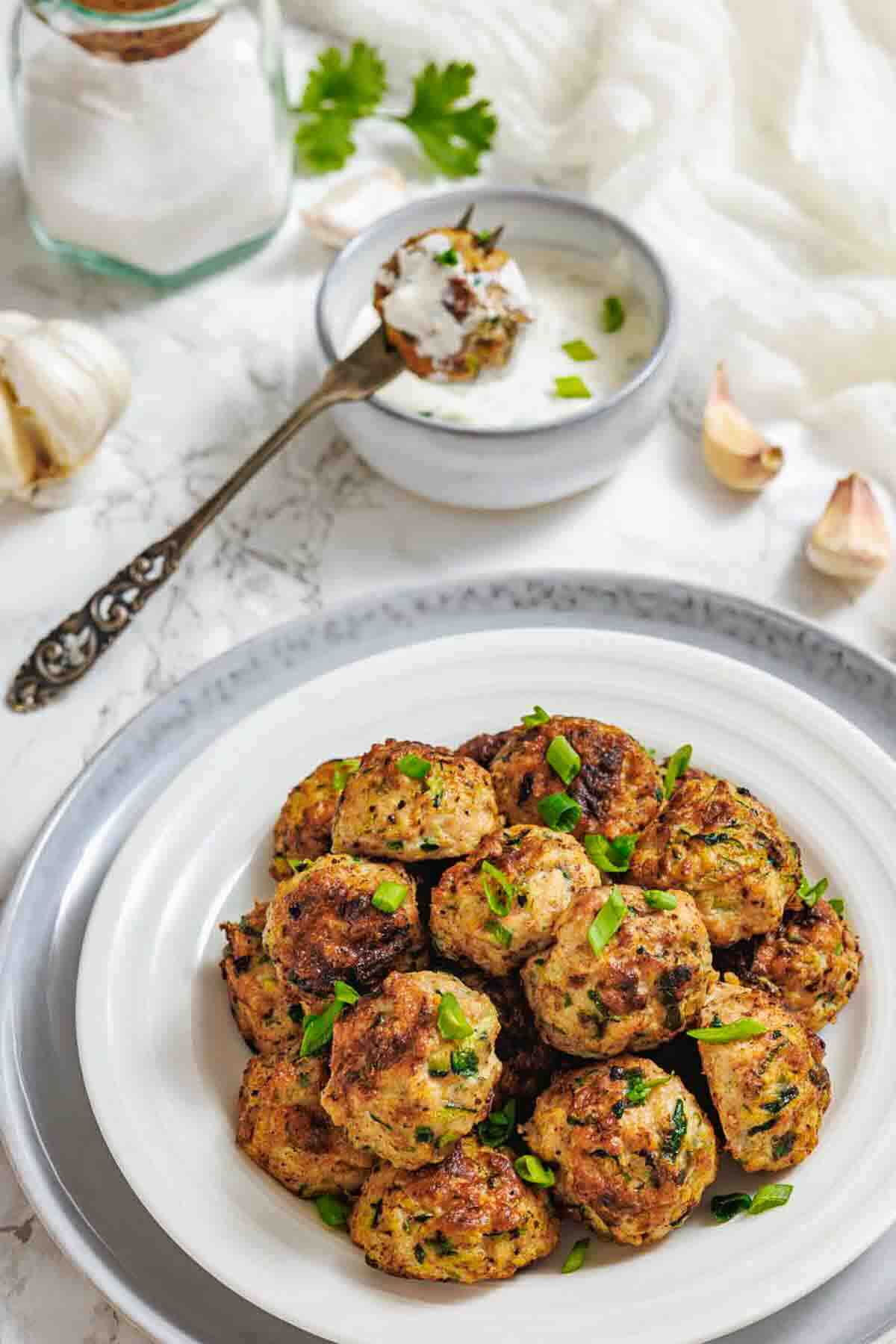 A white plate of browned meatballs garnished with chopped herbs, with a small bowl of creamy dipping sauce and a spoon in the background.