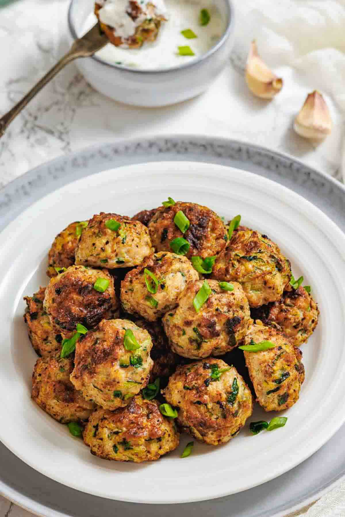 A white plate filled with herb-studded chicken meatballs, garnished with chopped green onions, with a bowl of dipping sauce and garlic cloves in the background.