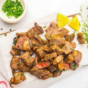 Cubed pieces of cooked steak garnished with chopped herbs, served with lemon wedges on a white surface, with a bowl of herbs in the background.