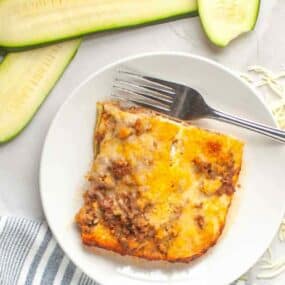 A slice of baked lasagna with ground meat and cheese on a white plate with a fork, next to sliced zucchini.