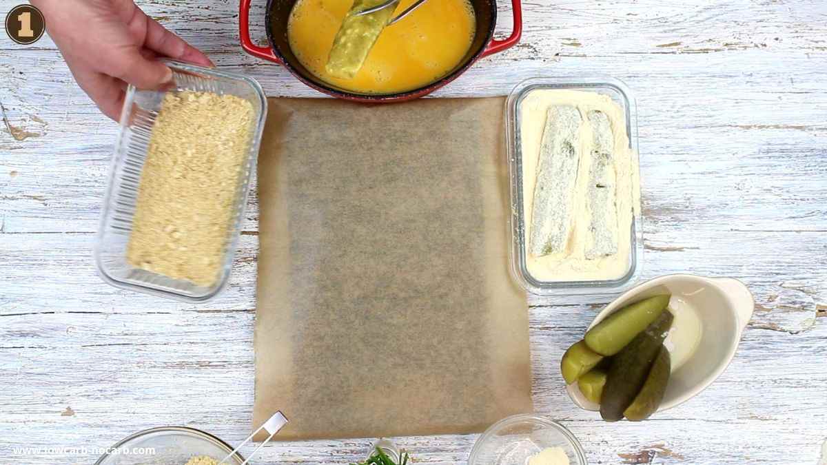 A hand dips pickle slices in breadcrumbs, with bowls of egg wash, flour, and sliced pickles on a white wooden surface.