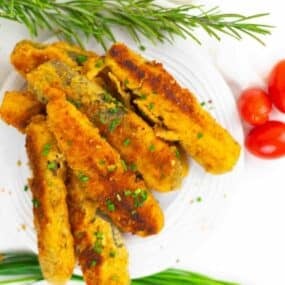 A plate of breaded and fried finger-shaped pieces garnished with chopped herbs, with sprigs of rosemary and cherry tomatoes beside the plate.