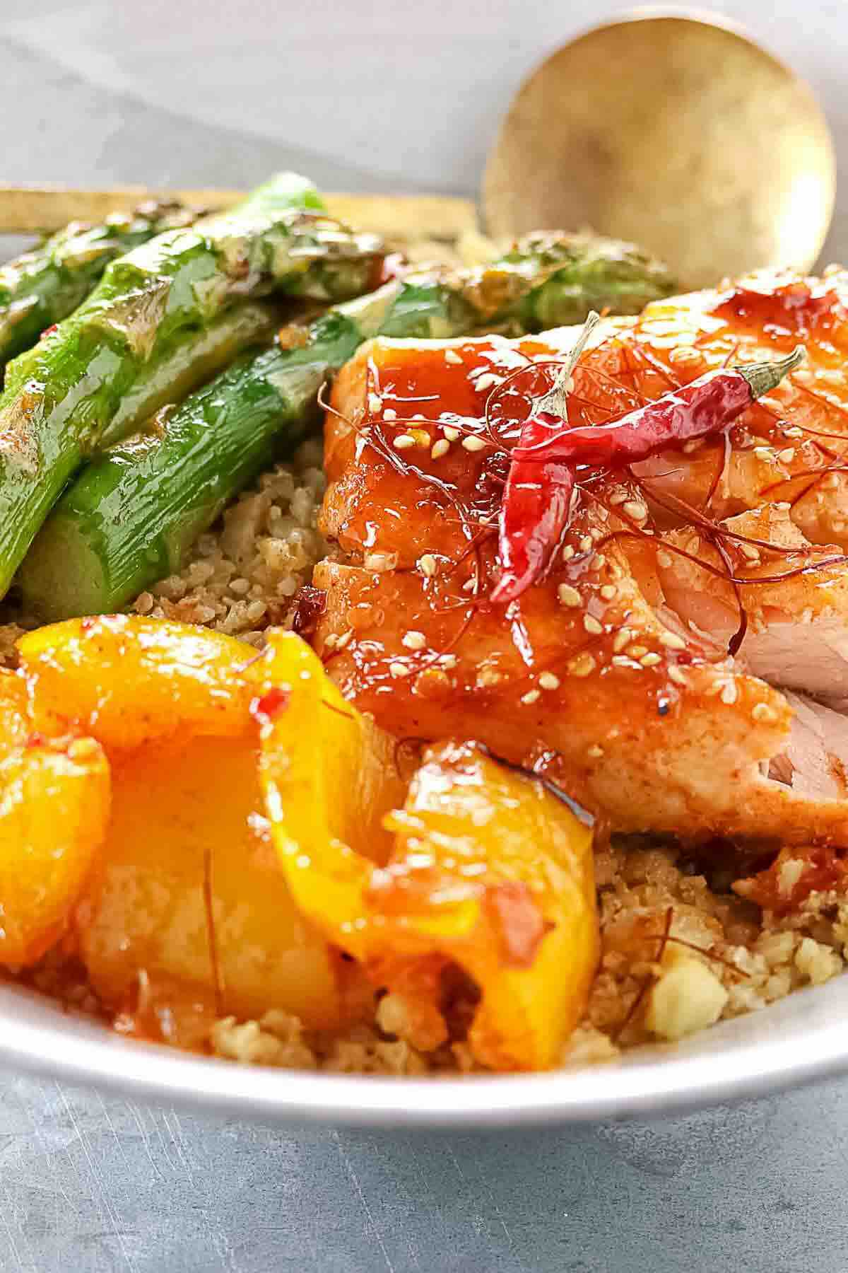 Bowl of quinoa topped with grilled chicken, asparagus, roasted yellow bell peppers, sesame seeds, and chili flakes, with a gold spoon in the background.