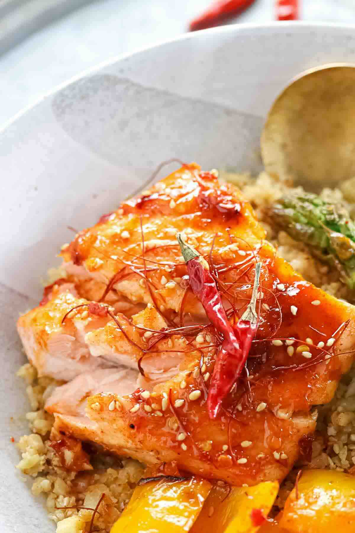 A plate of glazed salmon topped with sesame seeds, red chili, and shredded chili threads, served with quinoa, yellow bell pepper, and asparagus.