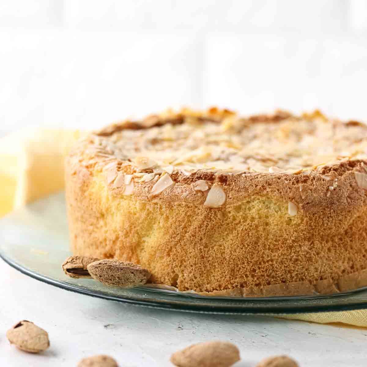 A round almond cake with a golden crust sits on a glass plate, with a few whole almonds scattered around the base.