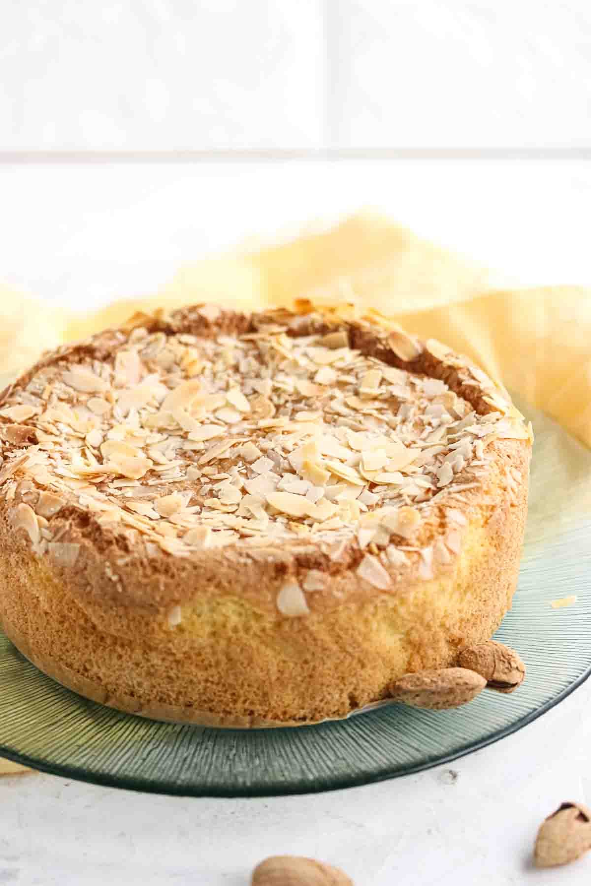 A round almond cake topped with sliced almonds sits on a green glass plate with a few whole almonds beside it.