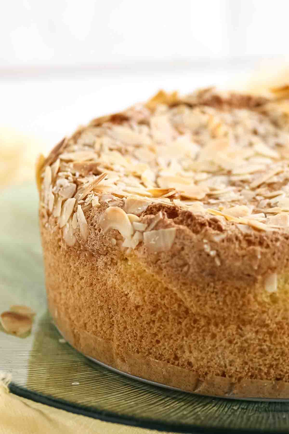 A close-up of a round sponge cake topped with sliced almonds, displayed on a glass plate.