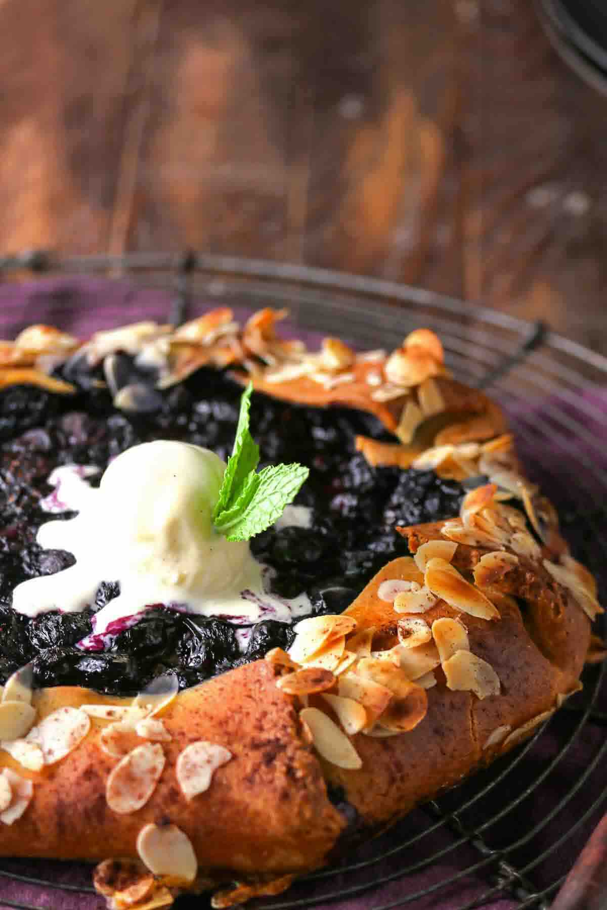 A rustic blueberry galette topped with a scoop of vanilla ice cream and a mint leaf, garnished with sliced almonds, sits on a wire rack.