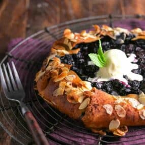 A rustic blueberry galette topped with a scoop of vanilla ice cream and a mint sprig, garnished with sliced almonds, sits on a wire rack next to a fork.